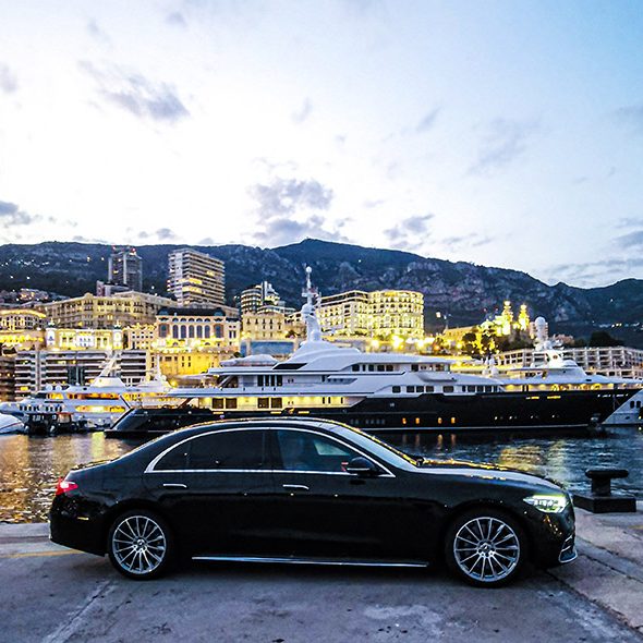 Black car on the harbor with an illuminated yacht and buildings in the background.