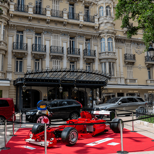 Red racing car displayed in front of an elegant building with balcony.