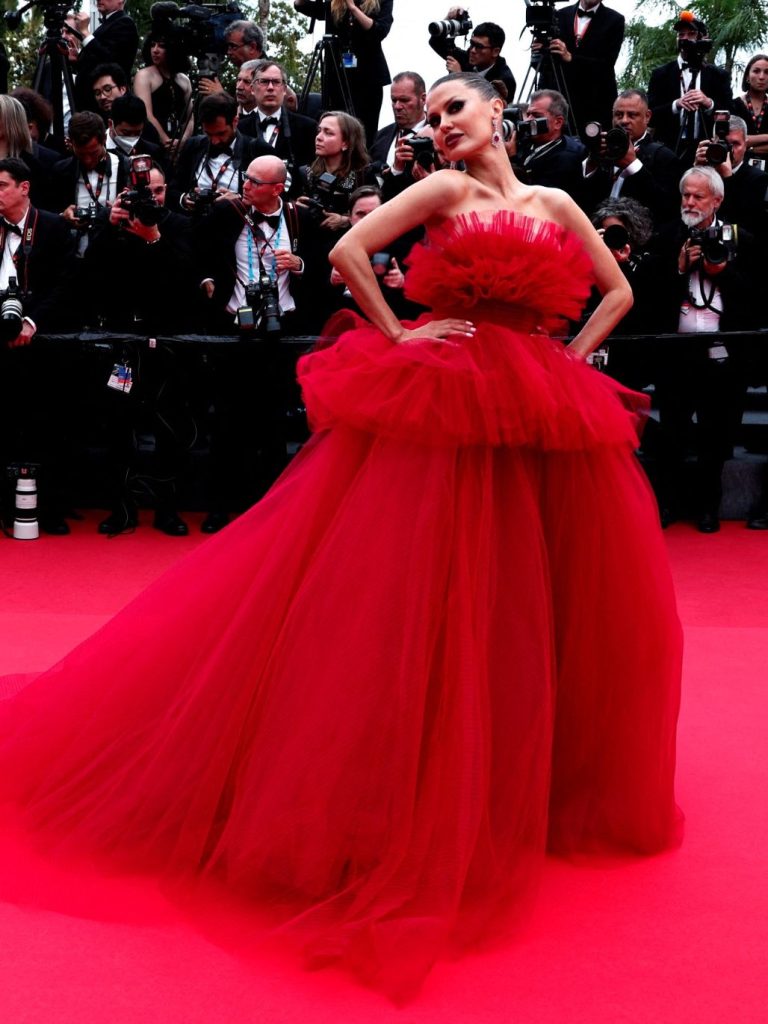 Woman in voluminous red dress on red carpet, surrounded by photographers.