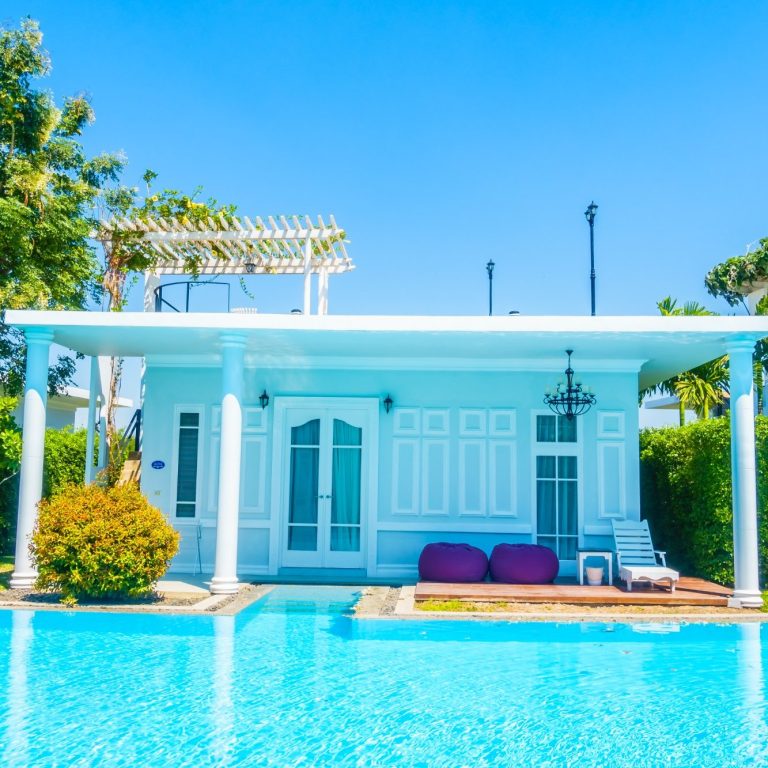 White villa with pool, lawn and comfortable chairs under a blue sky.