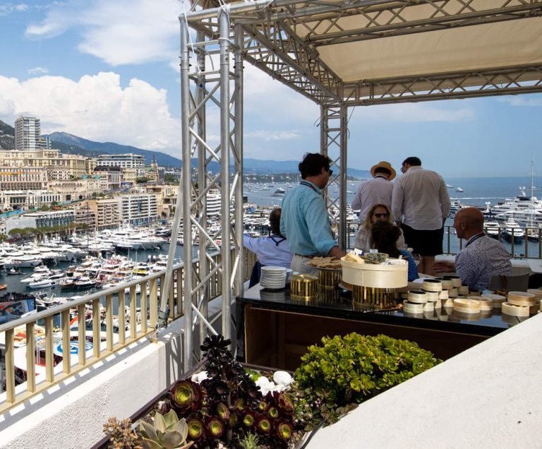 View of a terrace with people chatting and yachts in port in the background.