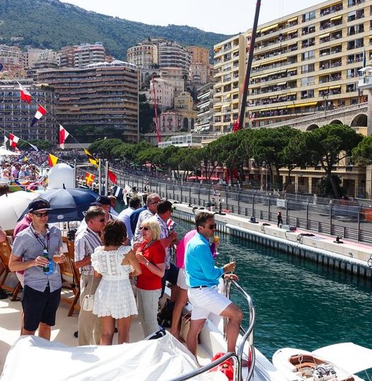 People on a yacht in Monaco, with buildings and a promenade in the background.