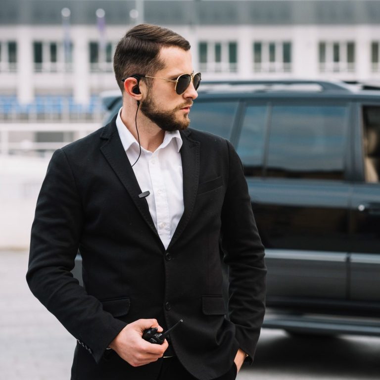 Man in black suit with sunglasses, listening to a walkie-talkie near a car.